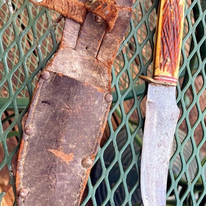 May include: A vintage hunting knife with a brown leather sheath. The knife has a brown and red handle with a metal blade.
