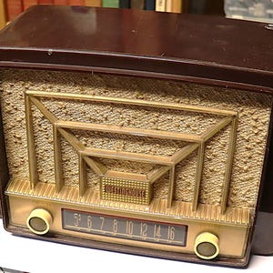 Puede incluir: Una radio Westinghouse vintage en una carcasa marrón oscuro. La radio presenta una rejilla de altavoz dorada y un dial con números. El logotipo de Westinghouse es visible. La radio tiene dos perillas color crema y una cubierta de altavoz texturizada.