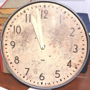 May include: Vintage round clock with a distressed, off-white face and black numerals. The clock has a metal frame and two cream-colored hands pointing to nearly 11 o'clock. The clock face has the letters "IBM" and the words "MADE IN U.S.A."