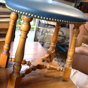 Op de afbeelding: Een vintage houten kruk met een ronde, blauwe leren zitting. De zitting is afgewerkt met messingkleurige studs. De poten zijn van gedraaid hout en de kruk heeft een klassieke, antieke uitstraling. Het hout heeft een warme, natuurlijke afwerking.