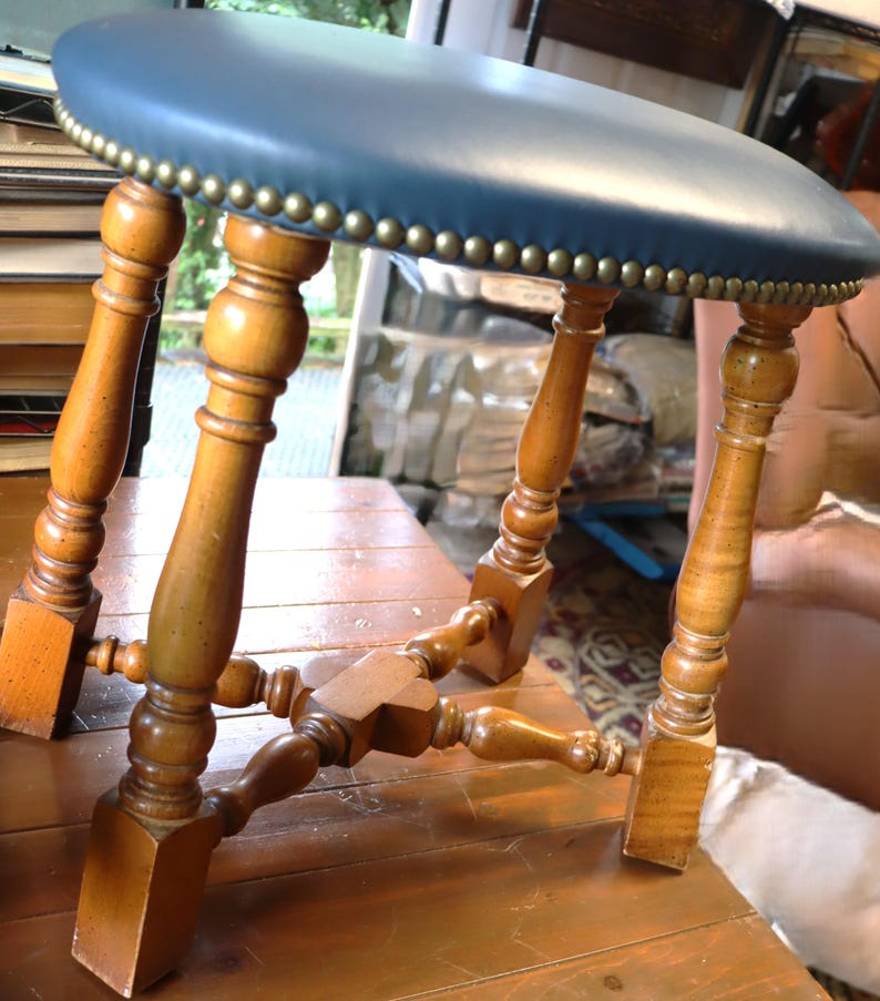 Op de afbeelding: Een vintage houten kruk met een ronde, blauwe leren zitting. De zitting is afgewerkt met messingkleurige studs. De poten zijn van gedraaid hout en de kruk heeft een klassieke, antieke uitstraling. Het hout heeft een warme, natuurlijke afwerking.