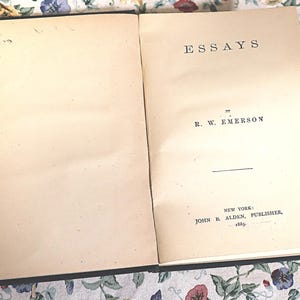 May include: Open book with the title "ESSAYS" by R. W. Emerson. The book is open to the title page, with the publisher's information at the bottom. The pages are aged with a cream color.
