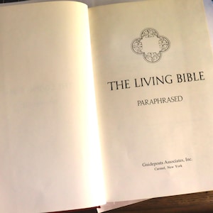 May include: Open book with the title "THE LIVING BIBLE" in black font. The word "PARAPHRASED" is below the title. A decorative emblem is at the top. The book is open to a cream-colored page.