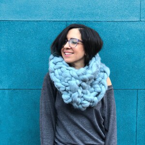 May include: A woman wearing a light blue chunky knit infinity scarf against a teal blue wall.