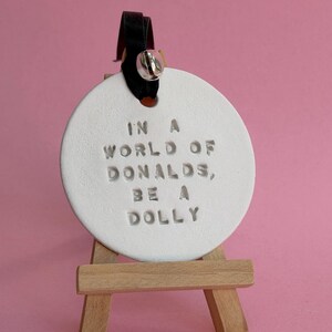 May include: A white circular ceramic ornament featuring the stamped text IN A WORLD OF DONALDS, BE A DOLLY. The ornament hangs from a black ribbon with a small silver bell and is displayed on a miniature wooden easel stand.