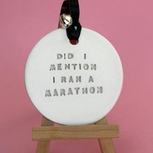 May include: A circular white ceramic ornament featuring the stamped text DID I MENTION I RAN A MARATHON. The ornament is suspended by a black ribbon with a small silver bell and is displayed on a miniature wooden easel stand.