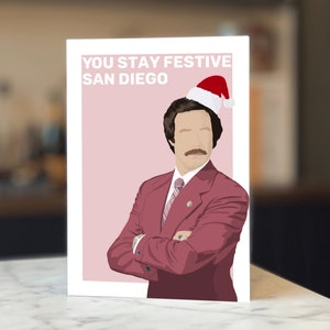 Op de afbeelding: Een wenskaart met een cartoon illustratie van een man in een rood pak en een kerstmuts. De kaart heeft de tekst "YOU STAY FESTIVE SAN DIEGO" in witte letters op een roze achtergrond. De kaart staat op een witte achtergrond.
