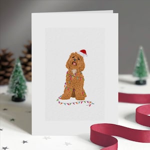May include: A white greeting card featuring a brown dog wearing a Santa hat and wrapped in colorful Christmas lights. The dog is sitting upright. The card is surrounded by Christmas decorations, including a red ribbon and miniature Christmas trees.