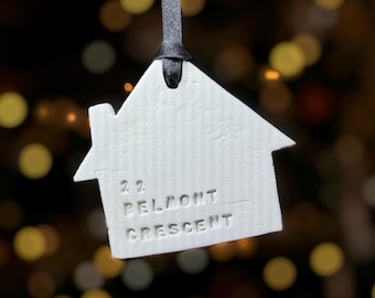 May include: A white ceramic house-shaped ornament with the address "22 BELMONT CRESCENT" embossed on the front. It hangs from a black satin ribbon. The background is blurred with warm, out-of-focus lights.