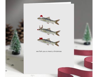 Op de afbeelding: Een witte wenskaart met drie vissen versierd voor de feestdagen. Een vis draagt een kerstmuts, een ander een gewei en de laatste een kroon. De tekst "we fish you a merry christmas" staat onderaan de kaart.