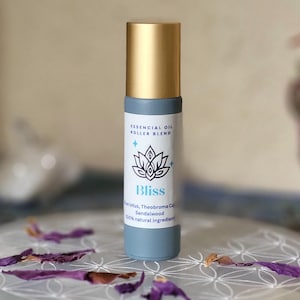 Könnte beinhalten: Eine blaue ätherische Öl-Roller-Mischung mit goldener Kappe. Das Etikett trägt die Aufschrift "Bliss" mit einem Lotusblumen-Design und listet die Inhaltsstoffe auf. Die Flasche ist von lila Blütenblättern auf einer weißen Oberfläche umgeben.