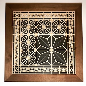 May include: A square wooden frame with a geometric pattern cut out of the center. The pattern is made of light wood and is set against a dark background. The frame is a dark brown wood.
