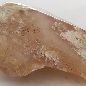 May include: A polished, irregularly shaped stone with a translucent, pinkish-tan hue. The stone has areas of creamy white and darker brown inclusions, creating a natural, textured appearance. The overall shape is elongated and smooth.