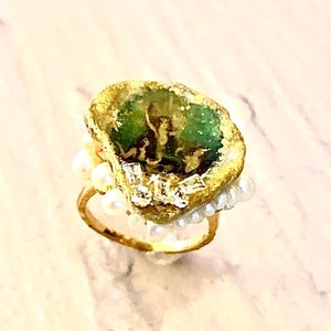 May include: A gold ring with a green stone set in a rough, irregular shape. The stone is surrounded by a border of small white pearls and clear crystals.