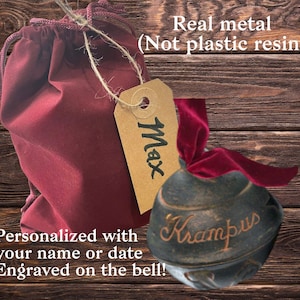 Op de afbeelding: Een donkerrode fluwelen tas met een bruin label waarop "Max" staat, ligt naast een donkerbruine metalen bel met het woord "Krampus" erop gegraveerd. De bel is vastgebonden met een donkerrood fluwelen lint. De achtergrond is een houten oppervlak.