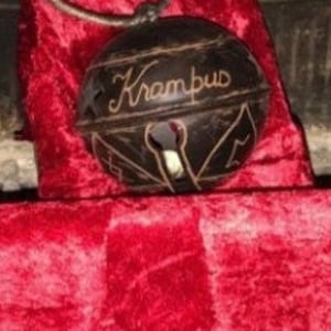 Può includere: Una campana marrone scuro con la parola "Krampus" in corsivo. La campana ha ritagli decorativi e poggia su una superficie di velluto rosso. La campana è sospesa da un cordino nero.