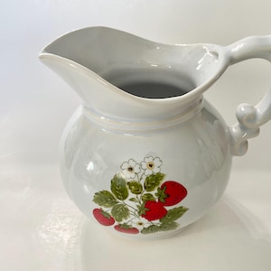 May include: White ceramic pitcher with a floral design featuring strawberries and white flowers. The pitcher has a round body and a handle.