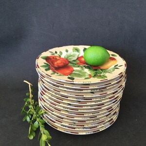 May include: A stack of 10 white ceramic dinner plates with a colorful fruit design. The plates feature a red apple, a green pear, and green leaves. A green lime sits on top of the stack.