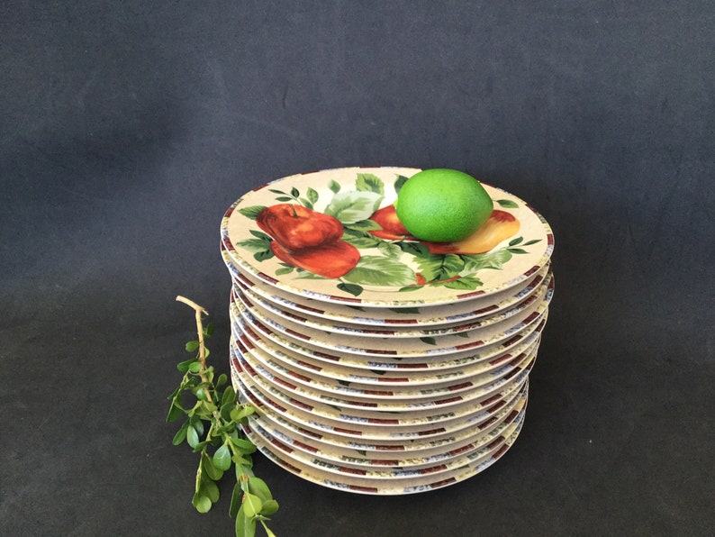 May include: A stack of 10 white ceramic dinner plates with a colorful fruit design. The plates feature a red apple, a green pear, and green leaves. A green lime sits on top of the stack.