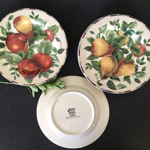 May include: Three ceramic dinner plates with a fruit design. The plates feature a white background with green leaves, red apples, and yellow pears. The plates have a brown rim with a decorative pattern. The back of one plate shows the words "Sakura Fresh Home" and "O'RCELL" in black.