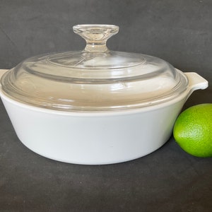 May include: White ceramic casserole dish with a clear glass lid. The dish has two handles on the sides. A green lime is sitting next to the dish.