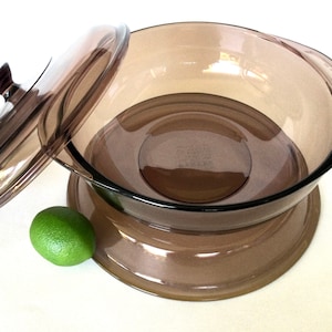 Op de afbeelding: Een bruine glazen serveerschaal met een bijpassend deksel en een verhoogde basis. De schaal heeft twee handvatten aan de zijkanten.