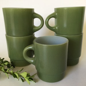 Puede incluir: Un conjunto de seis tazas apilables de color verde oliva. Las tazas tienen un asa redondeada y una parte superior ligeramente ensanchada. Las tazas están hechas de un material opaco y sólido y están dispuestas en grupo, algunas apiladas.