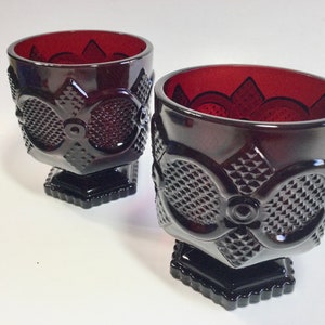 Op de afbeelding: Twee robijnrode kelkglazen met ingewikkelde reliëf bloemen- en geometrische patronen. De kelkglazen hebben een vierkante basis en een gedetailleerd ontwerp rond de kom. De binnenkant van de kelkglazen is dieprood.