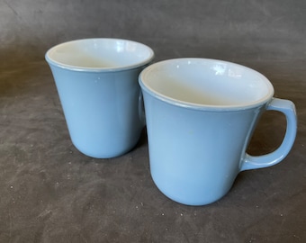 Może przedstawiać: Dwa niebieskie ceramiczne kubki z białym wnętrzem. Kubki są nieco różnej wielkości i mają zaokrąglony kształt.