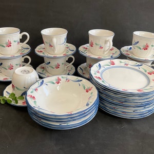 Op de afbeelding: Een set witte theekopjes en schoteltjes met blauwe en roze bloemenmotieven. De set bevat een stapel van 12 borden met een vergelijkbaar bloemenmotief.