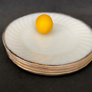 May include: A set of four white milk glass dinner plates with a gold rim. The plates are stacked on top of each other. A yellow lemon is sitting on top of the plates.