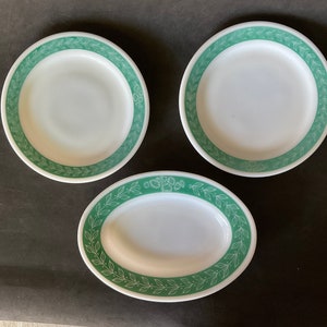 Könnte beinhalten: Drei weiße Keramikteller mit einem grünen Blattdesign am Rand. Die Teller liegen auf einer dunkelgrauen Oberfläche.