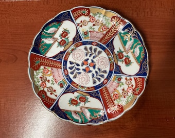 Può includere: Un piatto in ceramica bianca con un bordo ondulato e un design floreale colorato. Il piatto presenta un motivo centrale di fiori blu e rossi con accenti dorati, circondato da un motivo ripetitivo di fiori e foglie rossi, blu e verdi.