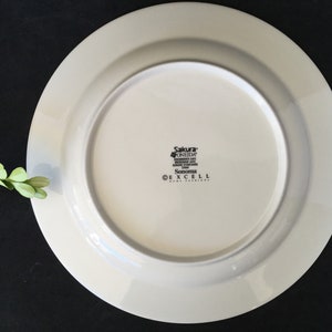 May include: White ceramic dinner plate with a raised rim. The bottom of the plate has the text "Sakura* Oneida, Dishwasher Safe, Microwave Safe, Oven Safe,  Sonoma, &copy; Excell Home Fashions".