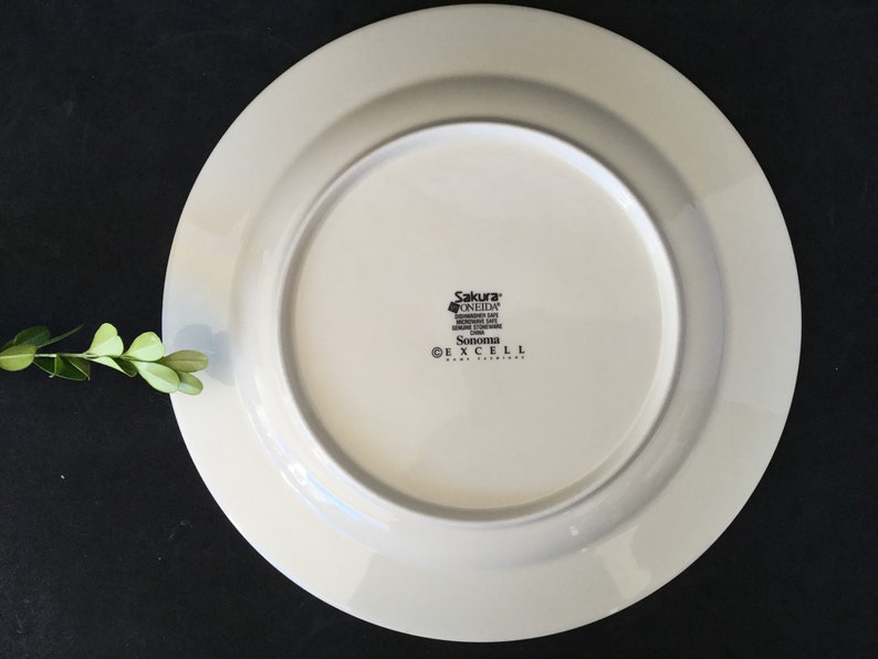 May include: White ceramic dinner plate with a raised rim. The bottom of the plate has the text "Sakura* Oneida, Dishwasher Safe, Microwave Safe, Oven Safe,  Sonoma, &copy; Excell Home Fashions".