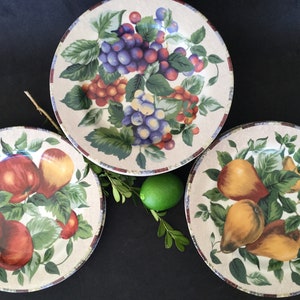 May include: Three ceramic dinner plates with a white background and colorful fruit designs. The plates feature grapes, apples, and pears.