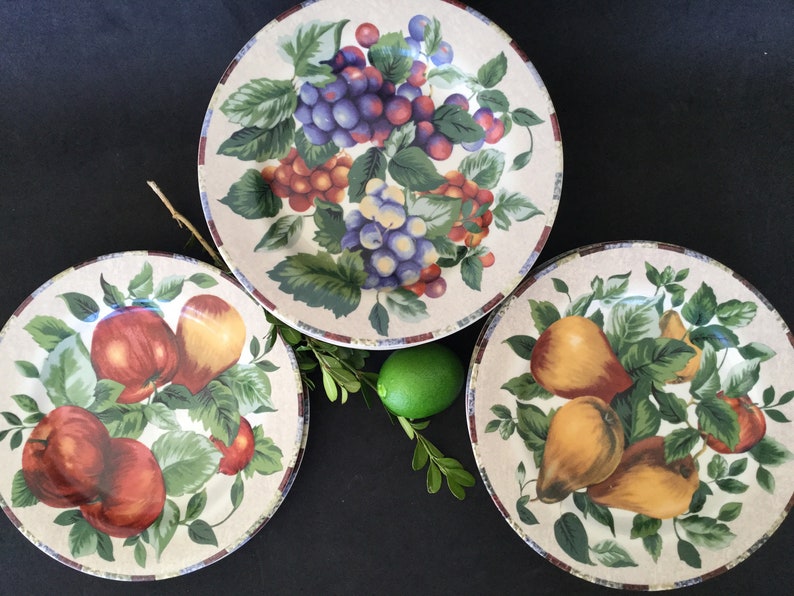 May include: Three ceramic dinner plates with a white background and colorful fruit designs. The plates feature grapes, apples, and pears.