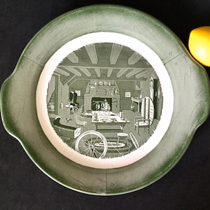 May include: A green ceramic serving platter with a white center featuring a black and white illustration of a rustic cabin interior. The platter has two handles on the sides.