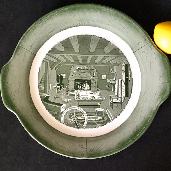 May include: A green ceramic serving platter with a white centre featuring a black and white illustration of a rustic cabin interior. The platter has two handles on the sides.