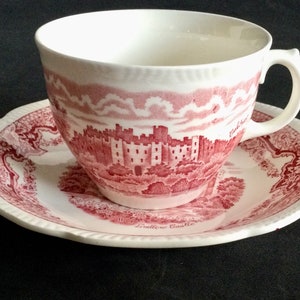 May include: A white teacup and saucer set with a red castle design. The teacup has a handle and sits on a matching saucer. The design includes the words "Luttrell Castle".