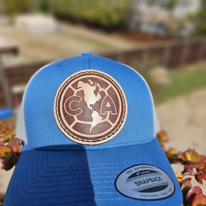 May include: A blue and white snapback hat with a brown leather patch featuring a circular design with the letters "CA" and a map of the Americas. The hat has a curved brim and a snap closure.