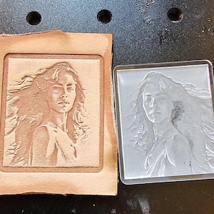 May include: A square, light brown leather piece with a carved portrait of a woman with flowing hair. Next to it is a clear, square acrylic piece with the same portrait etched into the surface. The image is on a dark surface.