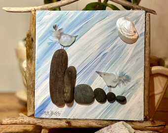 May include: A framed artwork featuring a coastal scene. The artwork depicts two birds made of glass and feathers, perched on stones. The background is painted with blue and white brushstrokes, and a seashell is attached. The frame is made of driftwood.