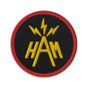 Pode incluir: Um remendo circular preto e vermelho com um design bordado amarelo. O design apresenta a palavra "HAM" com raios ao redor da letra "A".