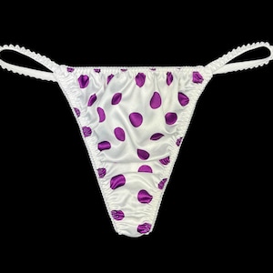 May include: White thong underwear featuring a purple polka dot pattern. The edges are trimmed with a scalloped design. The background is black.