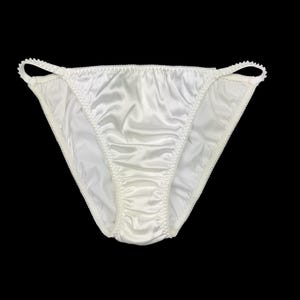 Puede incluir: Braga de satén blanco con un delicado ribete festoneado. La ropa interior tiene un corte de cintura baja y una textura suave y brillante. El fondo es negro.