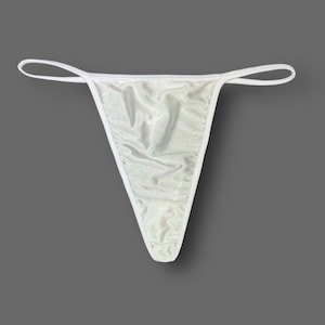 May include: White thong underwear with a shimmering, textured fabric. The thong features a triangular front and thin, white straps. The garment is displayed against a grey background.