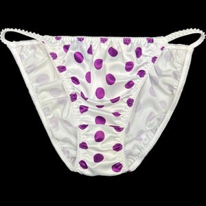 Può includere: Mutandine in raso bianco con motivo a pois viola. La biancheria intima a vita bassa ha un bordo arricciato e un delicato bordo smerlato. Lo sfondo è nero.