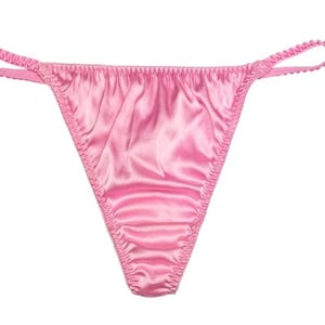 May include: A pair of light pink satin thong underwear. The thong features a low-rise waist and a narrow back. The edges are finished with a delicate, ruffled trim. The fabric has a subtle sheen.