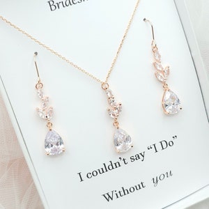 Puede incluir: Conjunto de collar y pendientes de oro rosa con un cristal en forma de lágrima y detalles de hojas. Las joyas se muestran en una caja blanca con el texto "No podría decir "Sí, quiero" Sin ti".